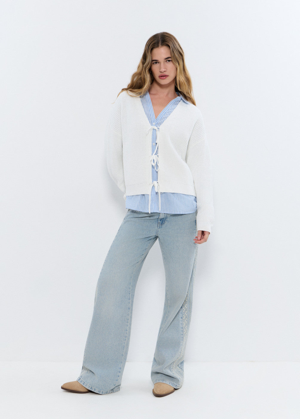 אישה לובשת קרדיגן לבן עם שרוכים, חולצה מכופתרת תכלת ומכנסי ג'ינס רחבים לאופנת נשים