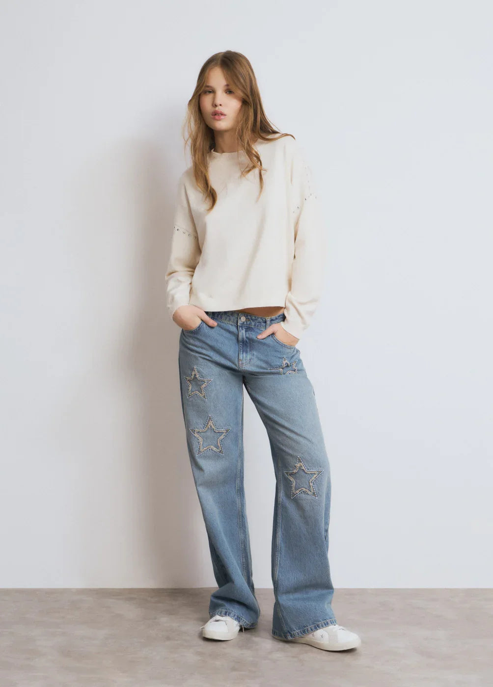 דוגמנית לובשת ג'ינס כותנה תכלת עם כוכבים ותפרים, סווטשירט קרם ונעלי סניקרס לבנות, אופנת נשים CHOZEN