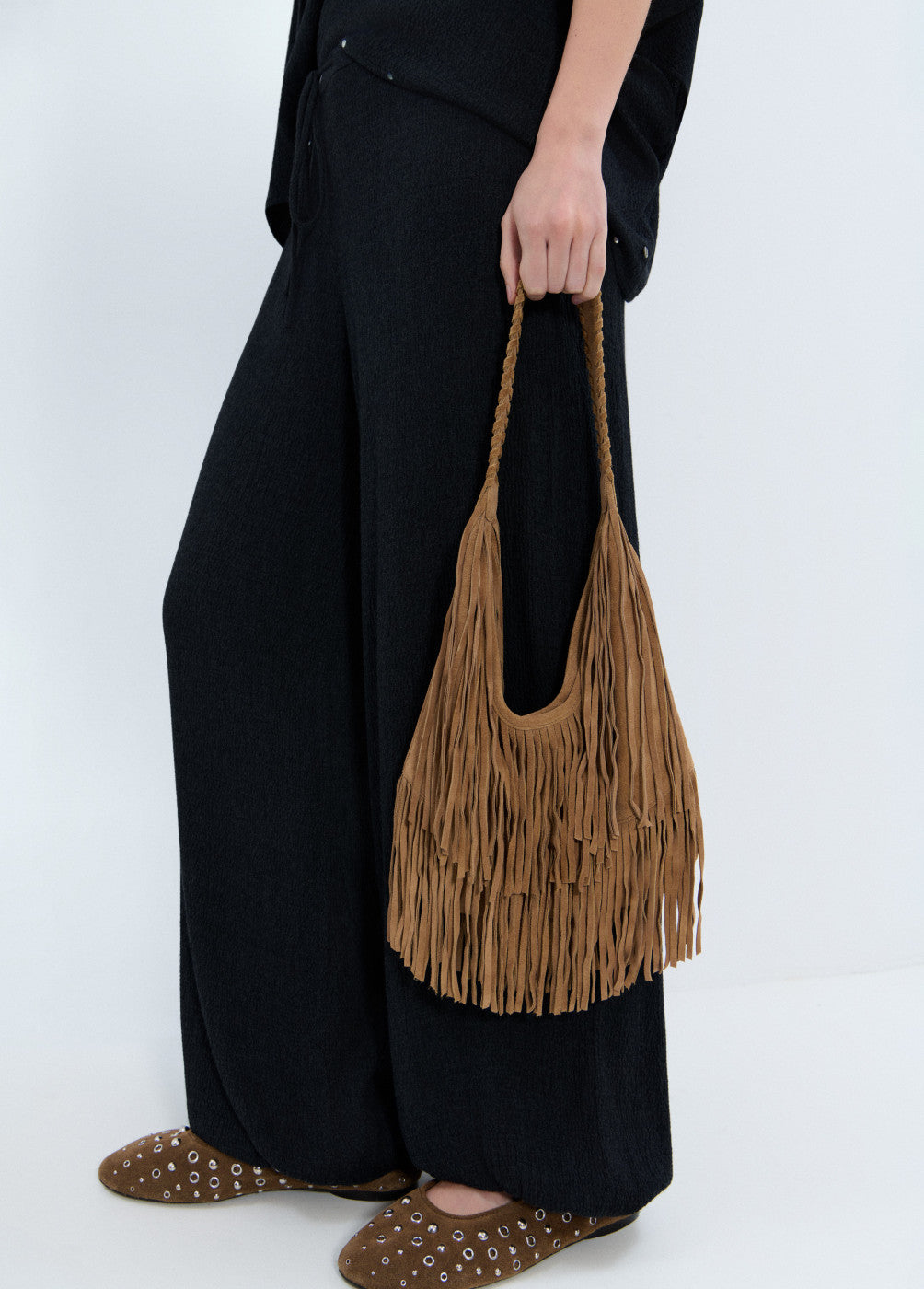 תיק צד חום עם פרנזים בעיצוב בוהו, מוחזק ביד אישה לבושה שחור ונעליים שטוחות חומות