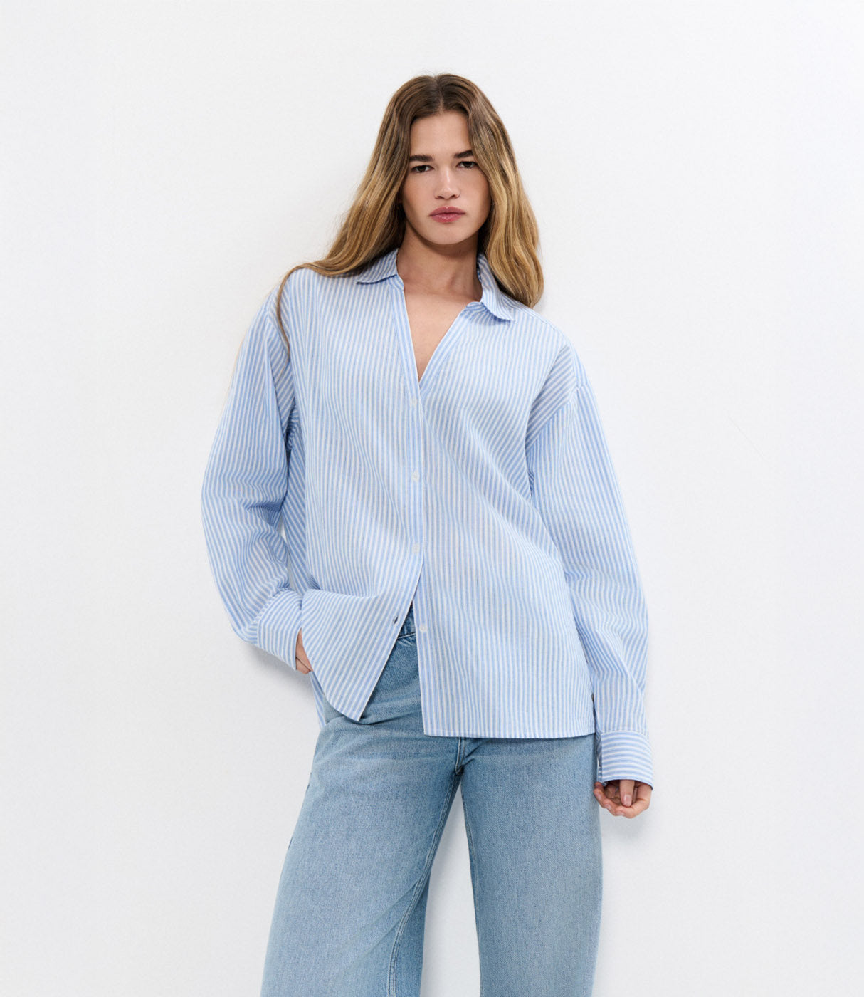 דוגמנית לובשת חולצת פסים כחול-לבן מכופתרת עם ג'ינס בהיר, רקע לבן