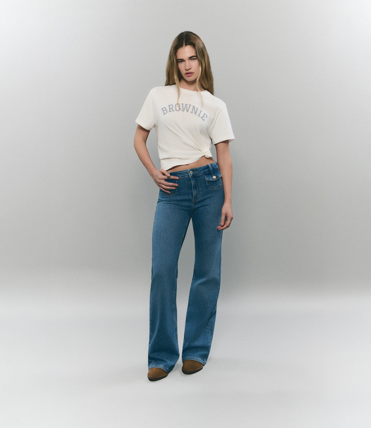אישה לובשת ג'ינס בגזרה ישרה וחולצה לבנה עם הדפס BROWNIE, רקע אחיד