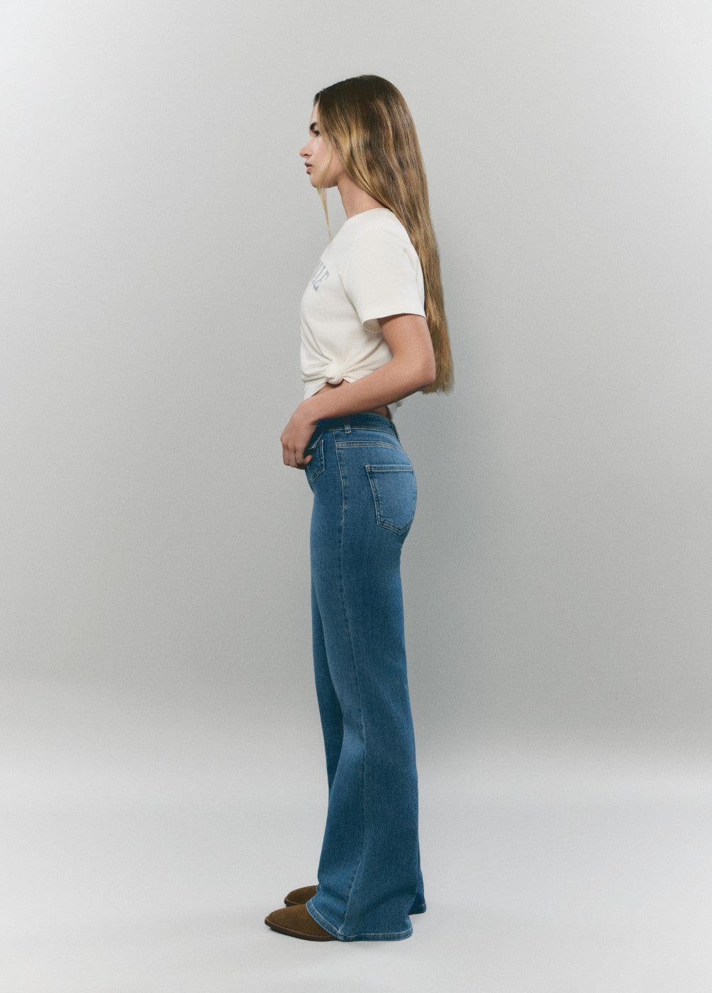 אישה לובשת ג'ינס בגזרת פדלפון וחולצה לבנה, צילום מהצד על רקע בהיר