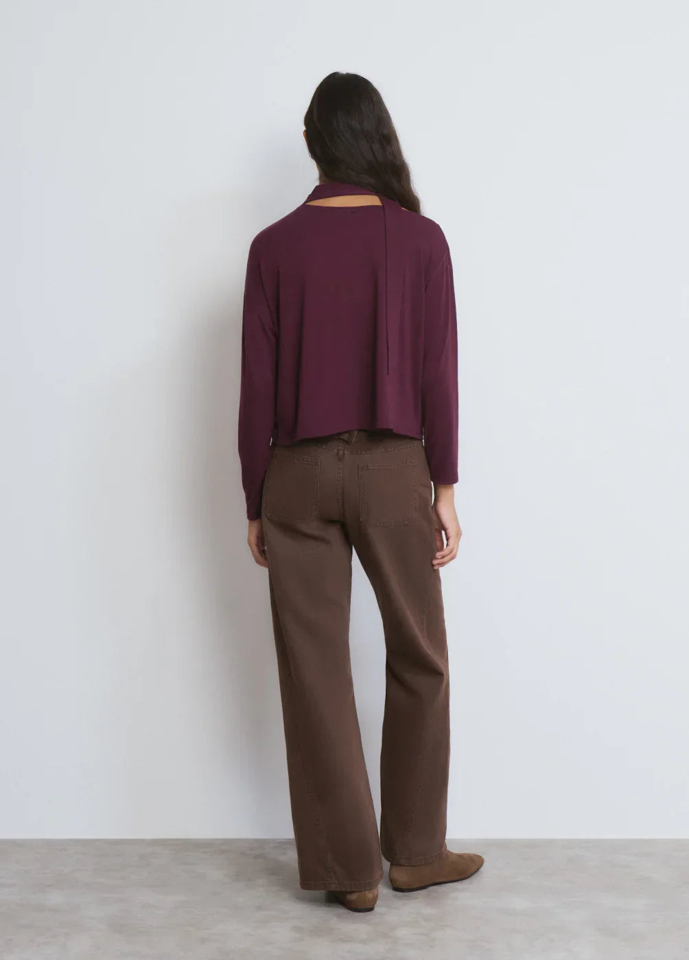 אישה עומדת עם גב למצלמה, לובשת חולצה בורדו ארוכה ומכנסיים חומים, צילום אופנה ל-CHOZEN