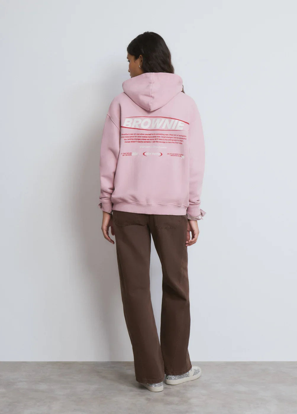 דוגמנית לובשת קפוצ׳ון ורוד עם הדפס BROWNIE וחולצת מכנסיים חומים בעמידה על רקע קיר לבן – אופנת נשים CHOZEN